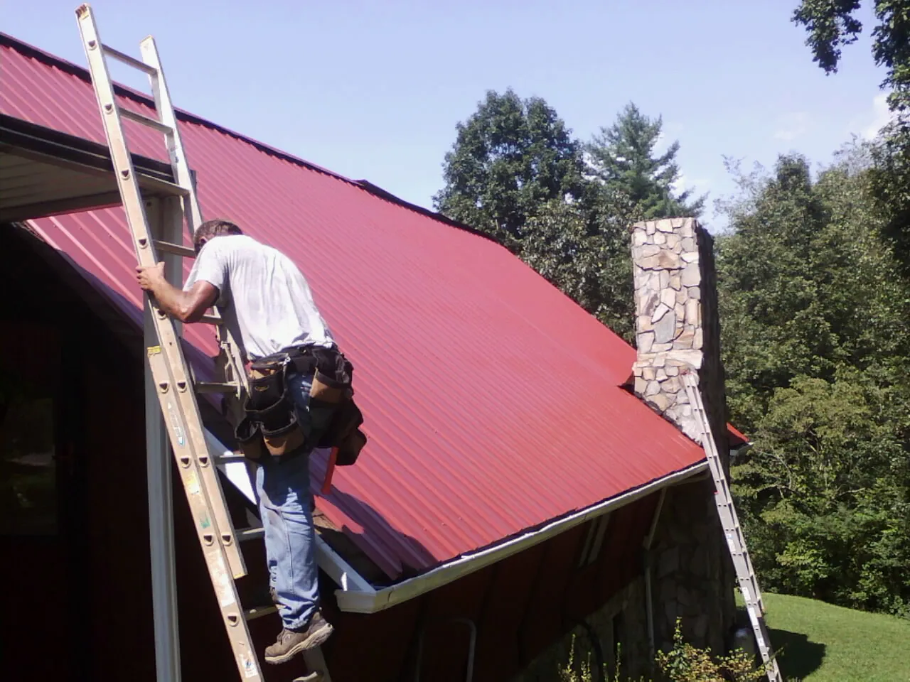 Roofer on a ladder beside a red metal roof and stone chimney — Soffit & Fascia Repair by Roofing Brothers in Horton